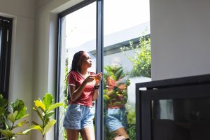A young woman enjoys a slice of pizza by a sunny window. She's dressed casually in a red top and denim shorts, reflecting a relaxed home setting.