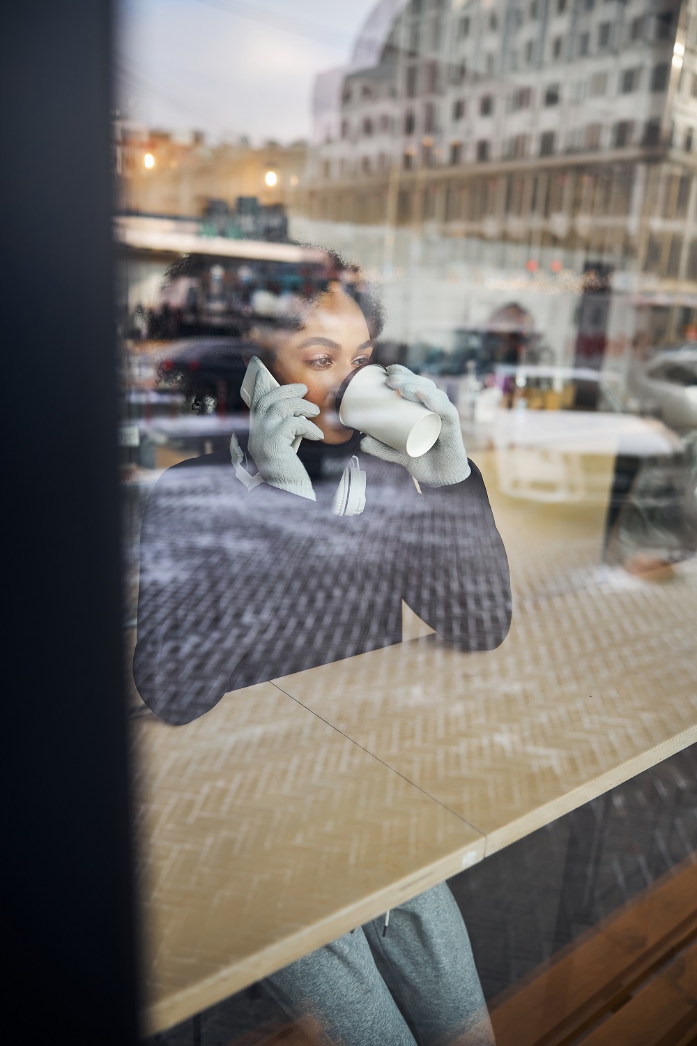 Person sitting at a window-side table, holding a smartphone to their ear and drinking from a paper cup, with city buildings reflected in Security and Safety Film on the glass.