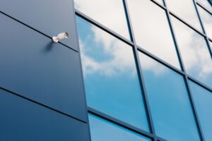 A security camera mounted on the exterior of a modern glass building featuring Security and Safety Film, with clouds reflected in the large windows.