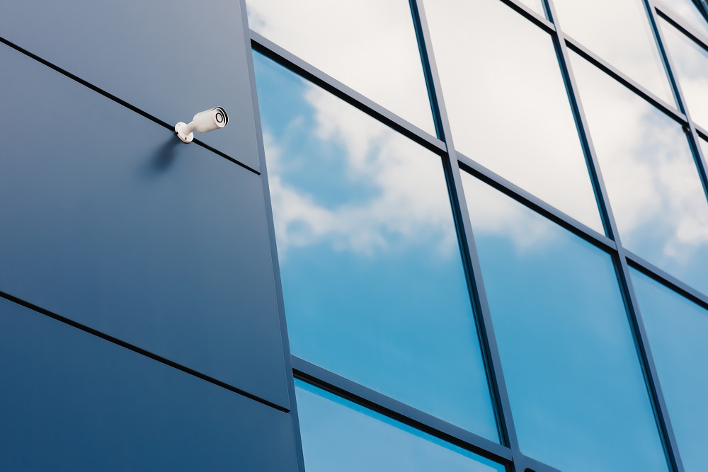 A security camera mounted on the exterior of a modern glass building featuring Security and Safety Film, with clouds reflected in the large windows.