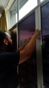 A man applies window tinting to a large sliding glass window in Slidell, with the glow of the sunset visible outside.