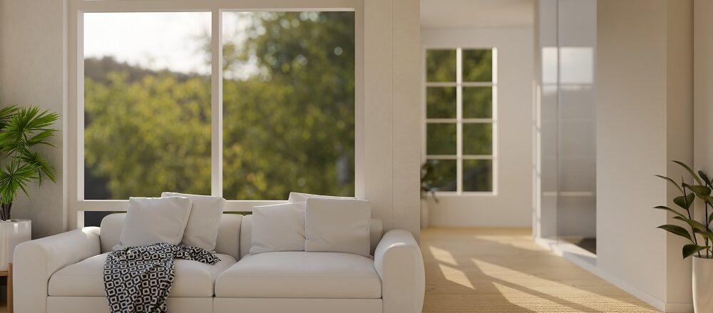 A modern living room with a white sofa, patterned throw blanket, large windows with Window Tinting St. Rose LA showing greenery outside, wooden floor, and potted plants.