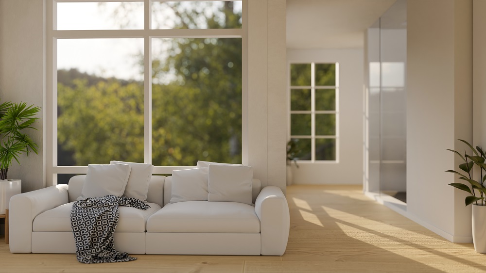 A modern living room with a white sofa, patterned throw blanket, large windows with Window Tinting St. Rose LA showing greenery outside, wooden floor, and potted plants.