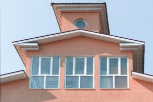The image shows the upper portion of a coral-colored building with white-framed windows, including one with window tinting in St. Rose, LA, and a small round window near the roof, set against a clear sky.